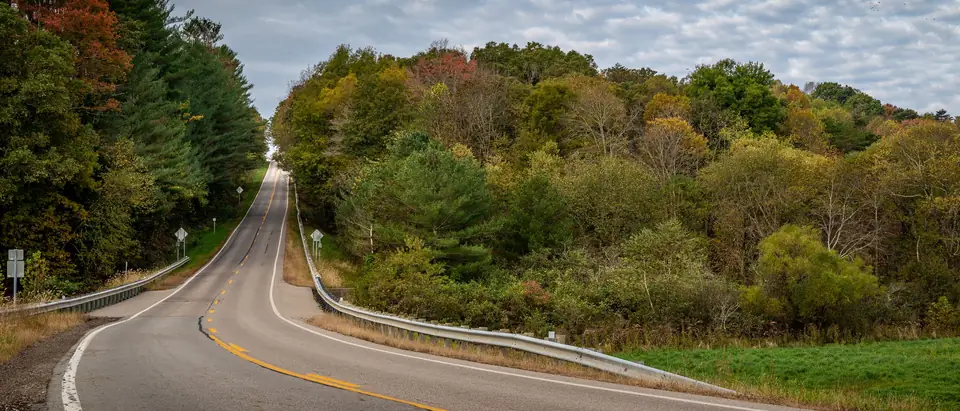 A two-lane road curving through autumn woods under a cloudy sky