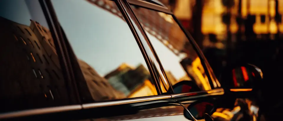 The side windows of a dark car reflecting buildings at sunset