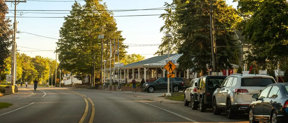 A small-town street with parked cars and tall trees in evening light